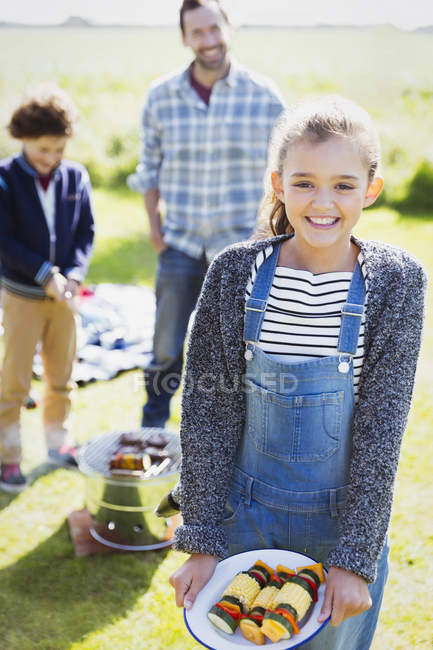 Retrato sorridente menina com espetos vegetais no acampamento ensolarado — Fotografia de Stock