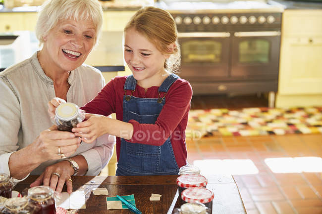 Abuela y nieta etiquetando frascos de conservas en la cocina - foto de stock