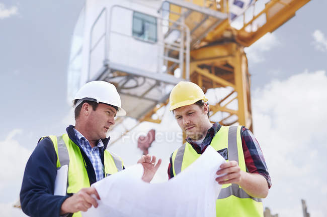 Construction worker and engineer reviewing blueprints below crane at construction site — Stock Photo