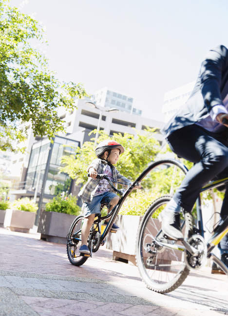 Figlio in casco in tandem in bicicletta con padre uomo d'affari nel soleggiato parco urbano — Foto stock