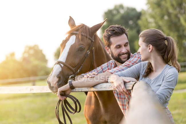 Pareja con caballo hablando en la cerca de pastos rurales - foto de stock