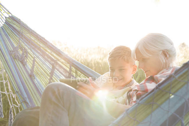 Бабушка и внук используют цифровые таблетки в солнечном гамаке — стоковое фото