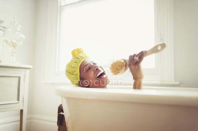 Playful mature woman singing into loofah brush in bathtub — Stock Photo