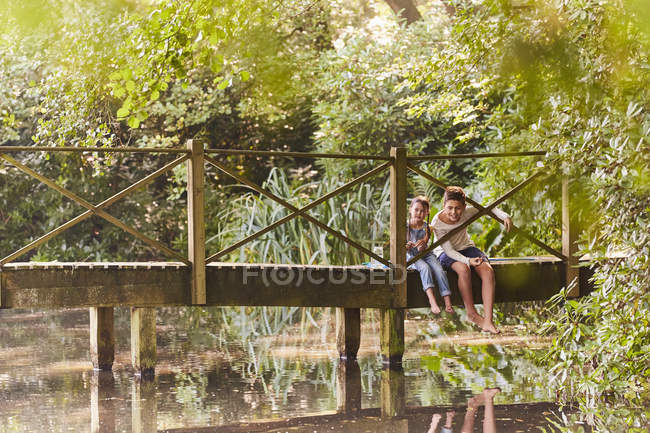 Fratello e sorella seduti sul ponte pedonale nel parco con alberi — Foto stock