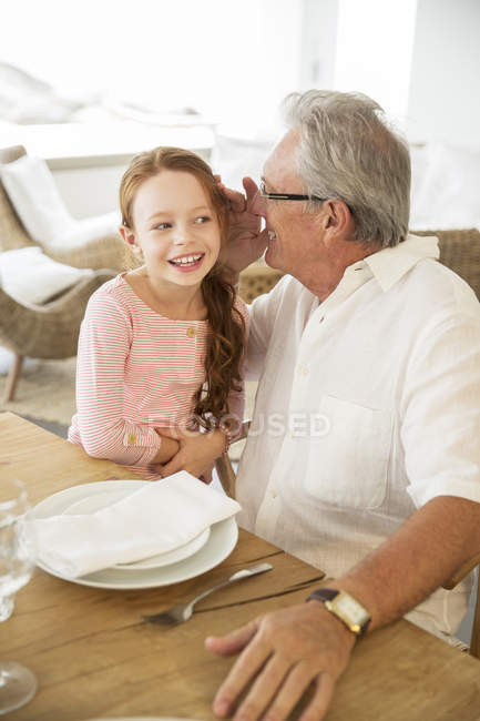 Older man and granddaughter whispering at table — Stock Photo