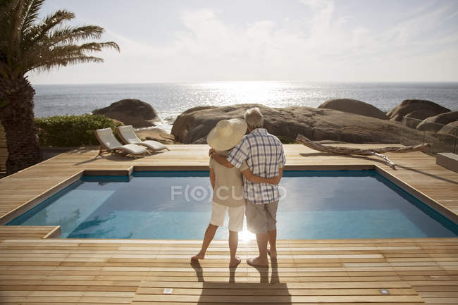 Couple sénior câlin par piscine moderne surplombant l'océan — Photo de stock