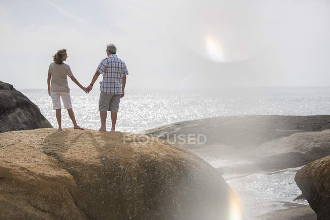 Старша пара тримає руки на скелях на пляжі — стокове фото