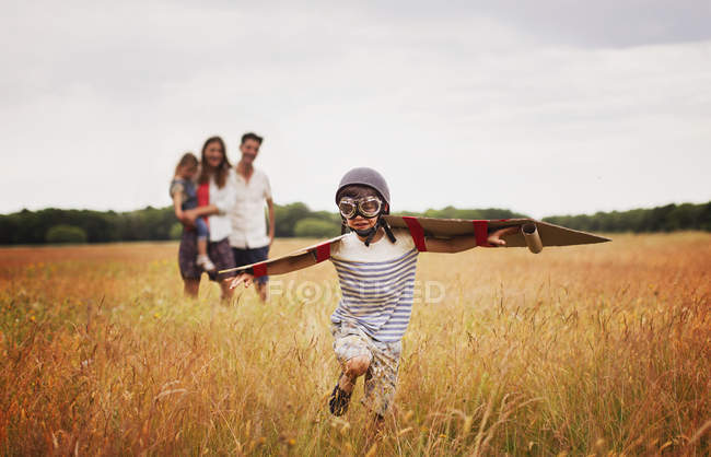 Playful boy with wings in aviators cap and flying goggles in field — Stock Photo
