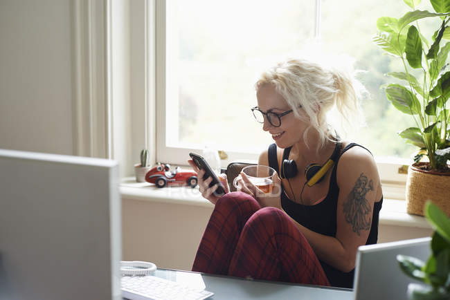 Mujer joven con auriculares y mensajes de texto de tatuaje en la oficina en casa - foto de stock