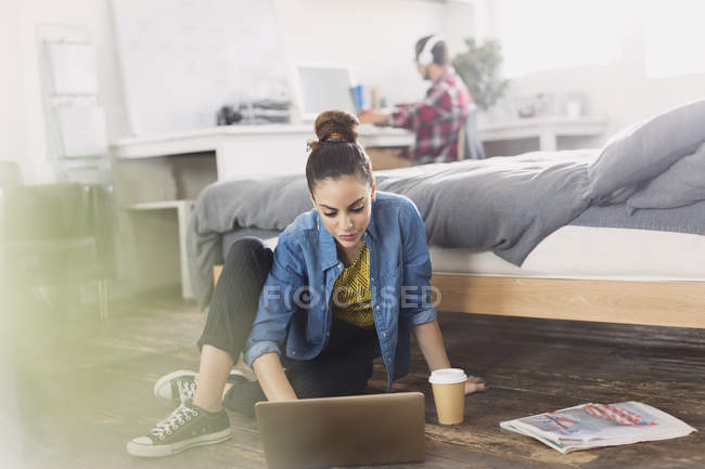 Female college student with coffee using laptop on bedroom floor — Stock Photo