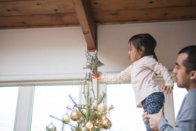 Father lifting toddler girl putting star on Christmas tree — Stock Photo