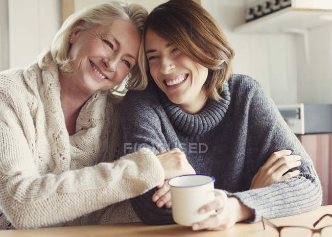 Смеющиеся мать и дочь в свитерах обнимаются и пьют кофе — стоковое фото