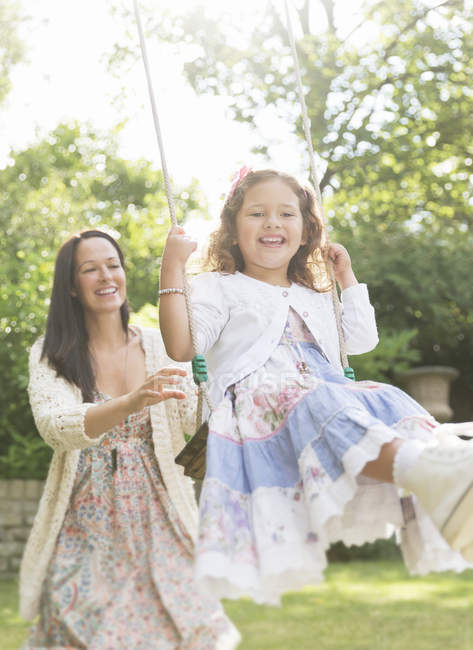 Mother in dress pushing daughter on swing in backyard — Stock Photo