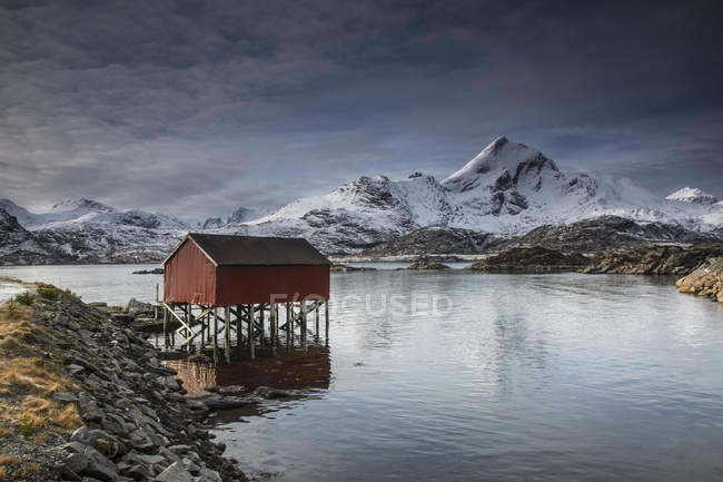 Montañas cubiertas de nieve detrás de la cabaña de pesca sobre el lago, Sund, Islas Lofoten, Noruega - foto de stock