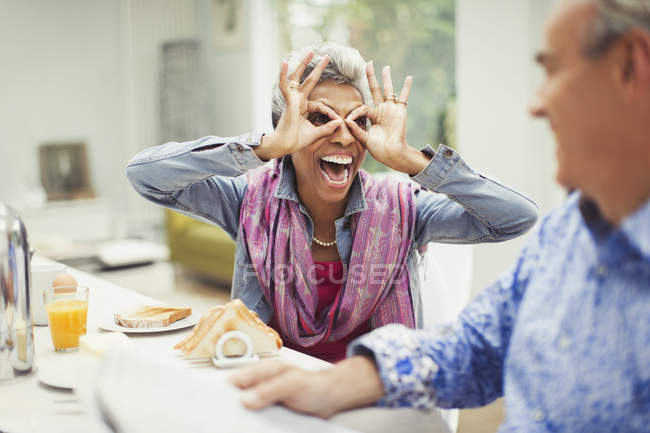 Verspielte reife Frau gestikuliert mit Fingergläsern am Frühstückstisch — Stockfoto