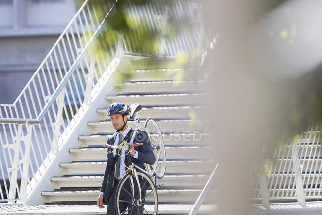 Empresario en traje y casco llevando bicicleta por escaleras urbanas - foto de stock