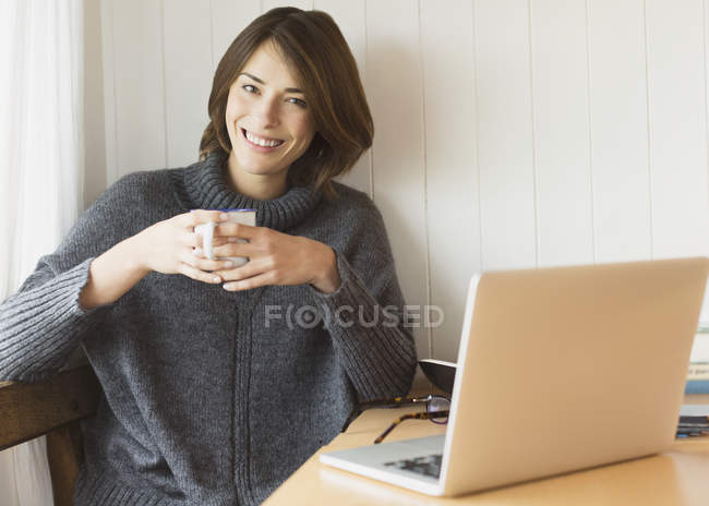 Ritratto sorridente donna bruna in maglione bere caffè al computer portatile — Foto stock