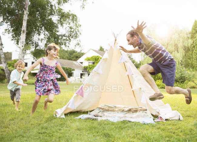 Father chasing children around teepee in backyard — Stock Photo