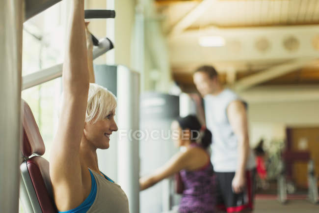 Lächelnde Frau mit erhobenen Armen an Trainingsgeräten im Fitnessstudio — Stockfoto