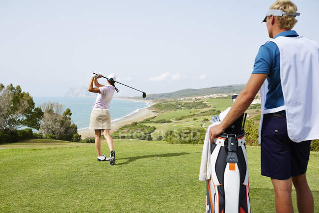 Caddy beobachtet Frau beim Abschlag auf Golfplatz mit Meerblick — Stockfoto