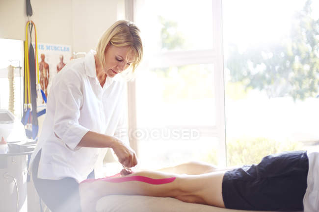 Fisioterapeuta aplicando cinta de kinesiología a la pierna del hombre - foto de stock