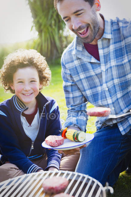 Father and son barbecuing hamburgers and vegetable skewers at grill — Stock Photo