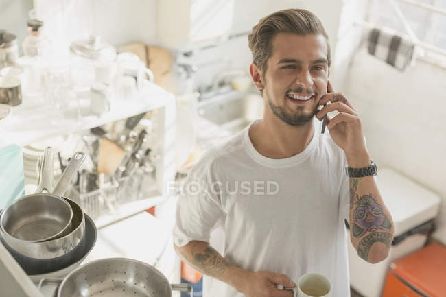 Tattooed young man talking on cell phone and drinking coffee in apartment kitchen — Stock Photo