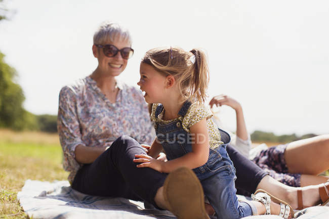 Grandmother and granddaughter laughing on blanket in sunny field — Stock Photo