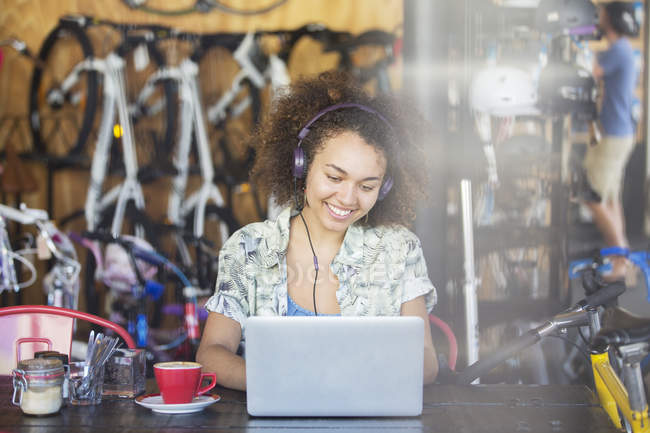 Smiling woman with headphones working at laptop in bicycle shop — Stock Photo