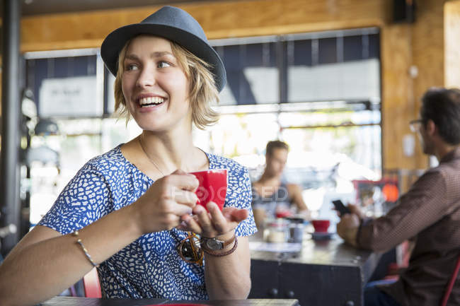 Mujer sonriente bebiendo café mirando por encima del hombro en la cafetería - foto de stock