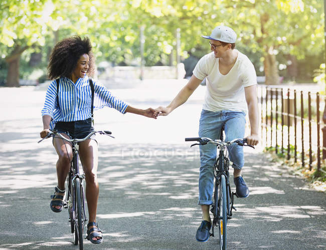 Coppia che si tiene per mano cavalcando biciclette nel parco urbano — Foto stock
