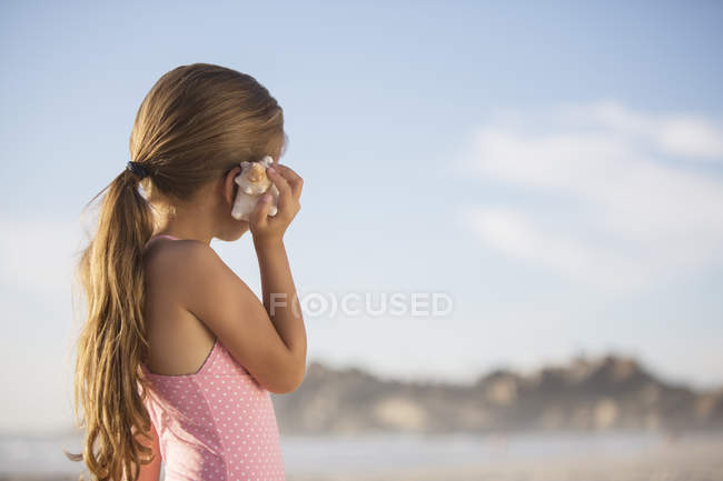 Chica escuchando concha marina en la playa - foto de stock