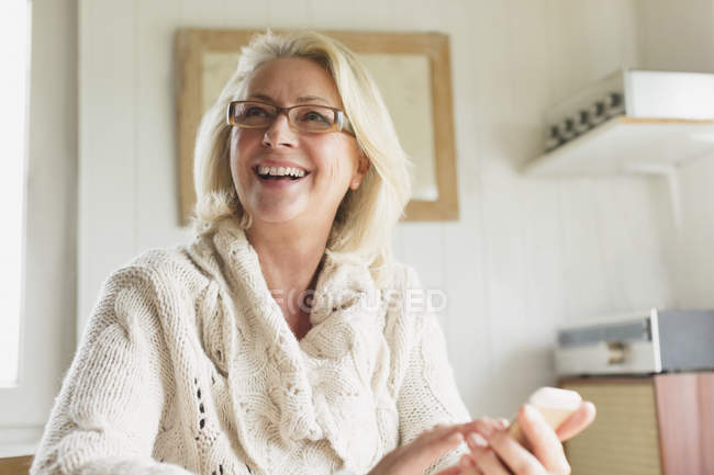 Sorrindo mulher idosa em camisola mensagens de texto com telefone celular na cozinha — Fotografia de Stock