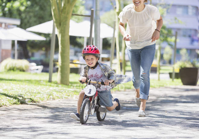 Madre persiguiendo hijo montar en bicicleta con casco en el parque soleado - foto de stock