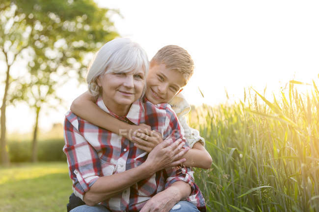 Портрет улыбающейся бабушки и внука, обнимающихся возле солнечного сельского пшеничного поля — стоковое фото