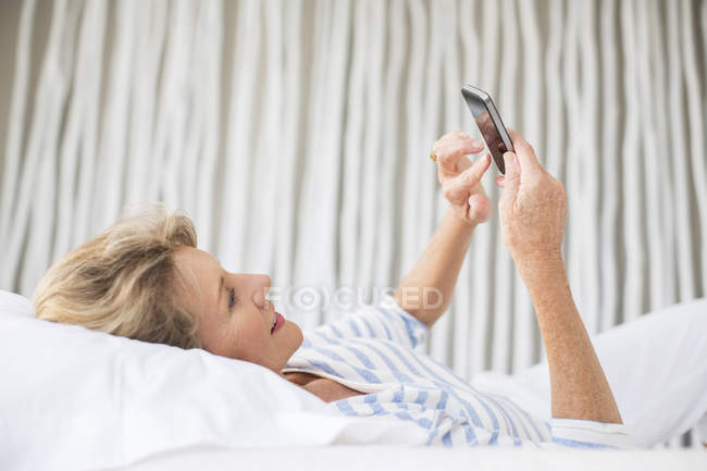 Літня жінка використовує мобільний телефон на ліжку — стокове фото