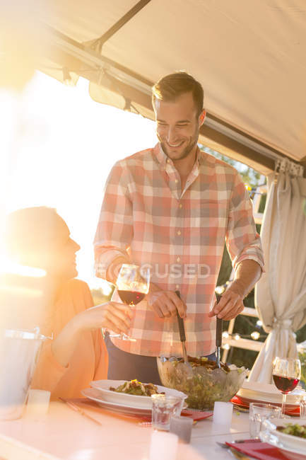 Young man serving salad to wife drinking wine at sunny patio table — Stock Photo