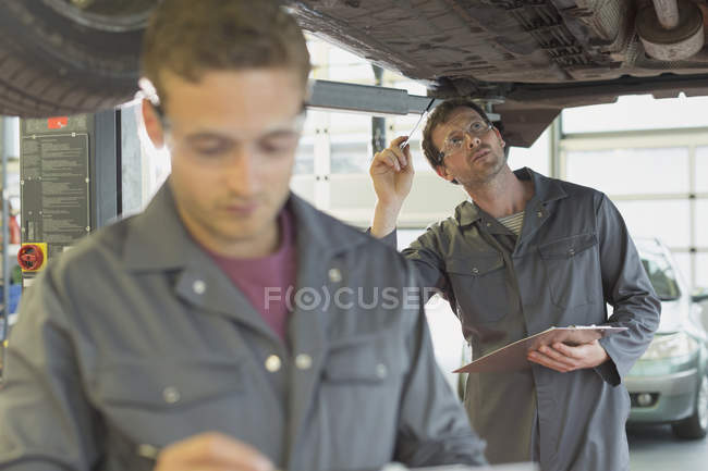 Mecânico com prancheta trabalhando sob o carro na oficina de reparação de automóveis — Fotografia de Stock