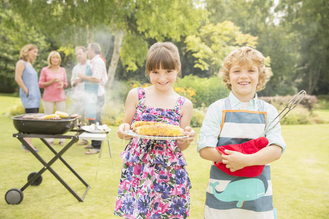 Brother and sister holding grilled corn near barbecue in backyard — Stock Photo