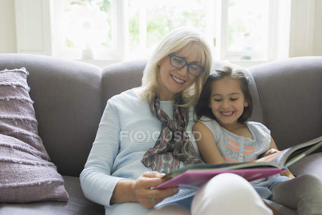 Grandmother and granddaughter reading book on living room sofa — Stock Photo