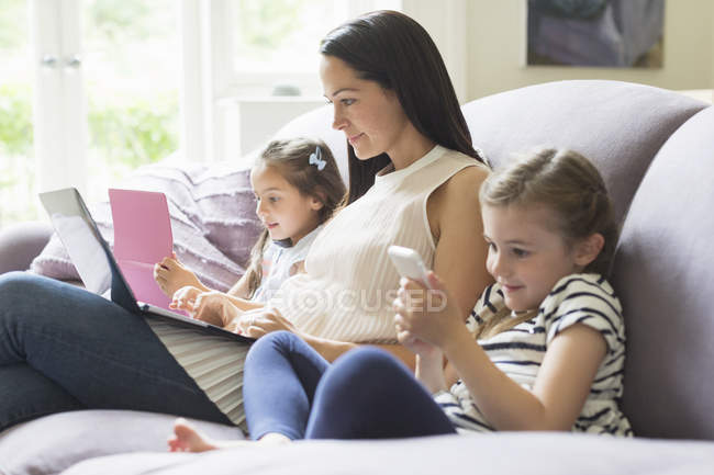 Mère et filles avec ordinateur portable, téléphone portable et tablette numérique sur canapé — Photo de stock
