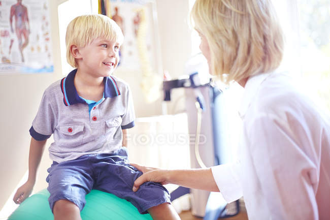 Физиотерапевт разговаривает с мальчиком на фитнес-мяч — стоковое фото
