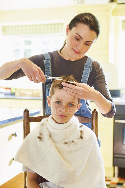 Portrait garçon malheureux se faire couper les cheveux de la mère dans la cuisine — Photo de stock
