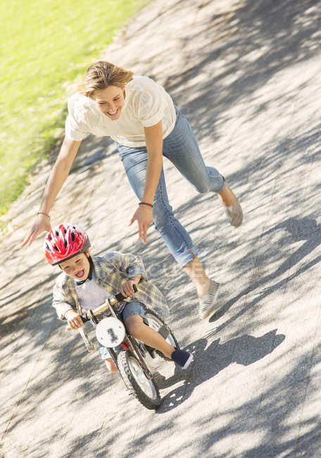 Mutter schubst Sohn mit Helm auf Fahrrad in sonnigem Park — Stockfoto