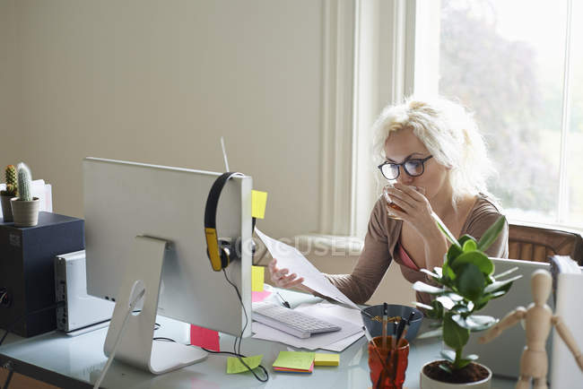 Молодая женщина пьет чай и читает бумажки на столе в домашнем офисе — стоковое фото