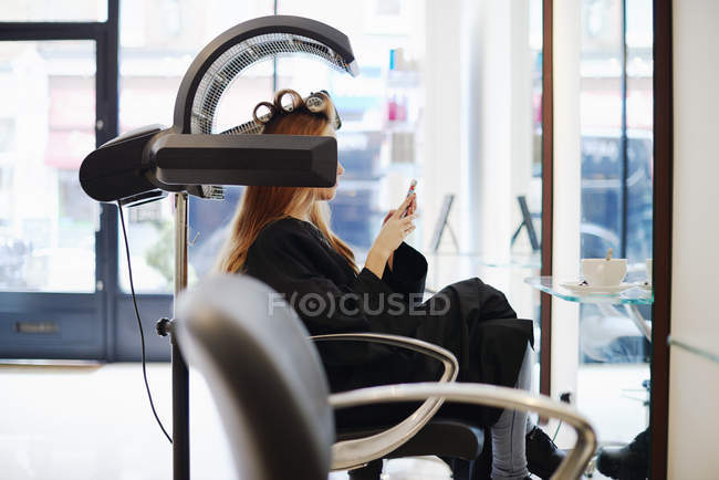 Femme assise sous sèche-linge textos avec téléphone portable dans le salon de coiffure — Photo de stock