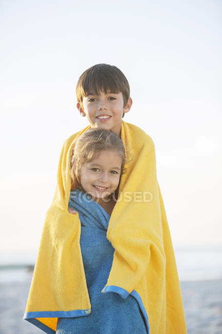 Hermano y hermana envueltos en toallas en la playa - foto de stock