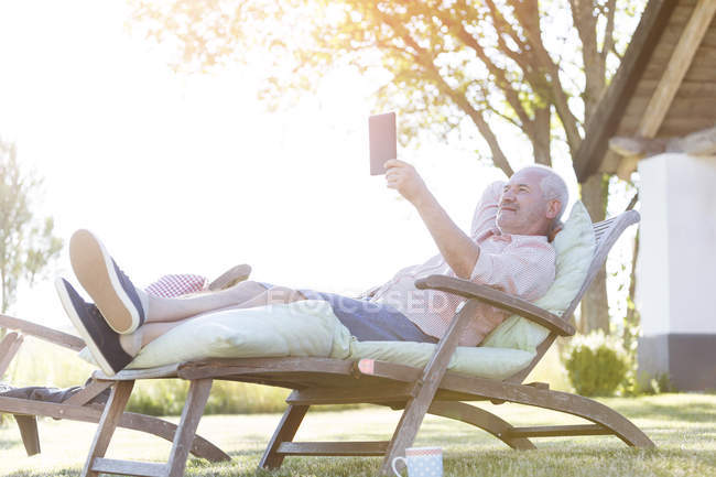 Homme âgé utilisant une tablette numérique relaxant sur une chaise longue dans la cour arrière — Photo de stock