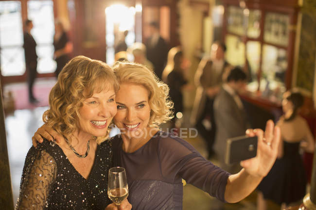 Mulheres bem vestidas com champanhe tomando auto-retrato com telefone câmera no lobby do teatro — Fotografia de Stock
