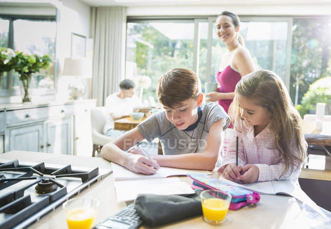 Mother watching brother and sister doing homework at kitchen counter — Stock Photo
